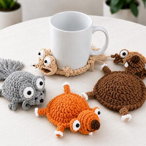 May include: Four crocheted coasters shaped like cartoon creatures in grey, tan, orange, and brown. Each coaster features large white eyes with black pupils, small limbs, and distinct facial expressions. One tan coaster is wrapped around a white mug.