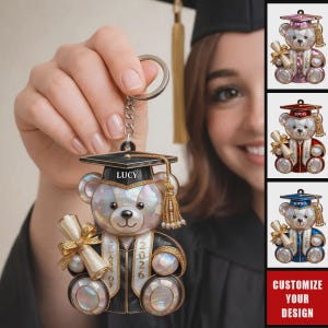 May include: A graduation-themed keychain featuring a bear wearing a graduation cap and gown, holding a diploma. The bear is adorned with iridescent accents and the text "LUCY" on the cap. Additional keychains with different names and colors are also shown.