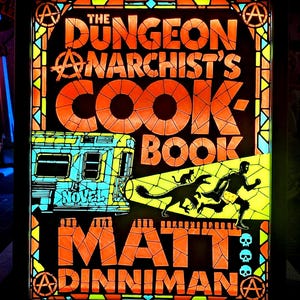 May include: A rectangular light box featuring a stained glass style design with the text THE DUNGEON ANARCHIST'S COOKBOOK NOVEL MATT DINNIMAN. The image displays an orange bus, a black silhouette of a person running with a dinosaur and cat.