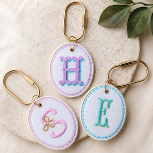 May include: Oval-shaped embroidered keychains with gold-colored hardware. Each keychain features a different initial: "H" in purple and blue, "S" in pink, and "E" in blue and green. The initials are embroidered on a white background.