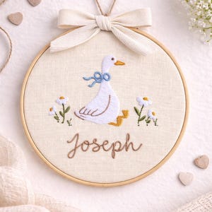 May include: A circular wooden embroidery hoop containing a cream-colored fabric panel. The fabric features an embroidered white goose with a blue bow, yellow feet, and an orange beak, flanked by small white flowers. The name Joseph is stitched below.