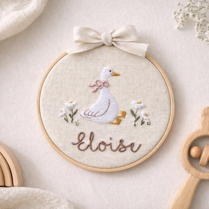 Könnte beinhalten: Gesticktes Kinderzimmer-Hängekunstwerk mit einer weißen Gans mit rosa Schleife, umgeben von gestickten Blumen. Der Name "Eloise" ist darunter gestickt. Ein Holzregenbogen und eine Bär-Rassel sind ebenfalls im Bild.
