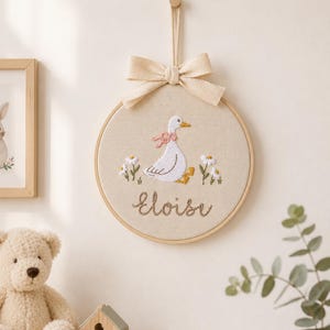 May include: A circular wooden embroidery hoop featuring a white goose with a pink neck ribbon, surrounded by small white flowers and green stems. The name Eloise is embroidered in brown script below the goose. A beige fabric bow hangs from the top.
