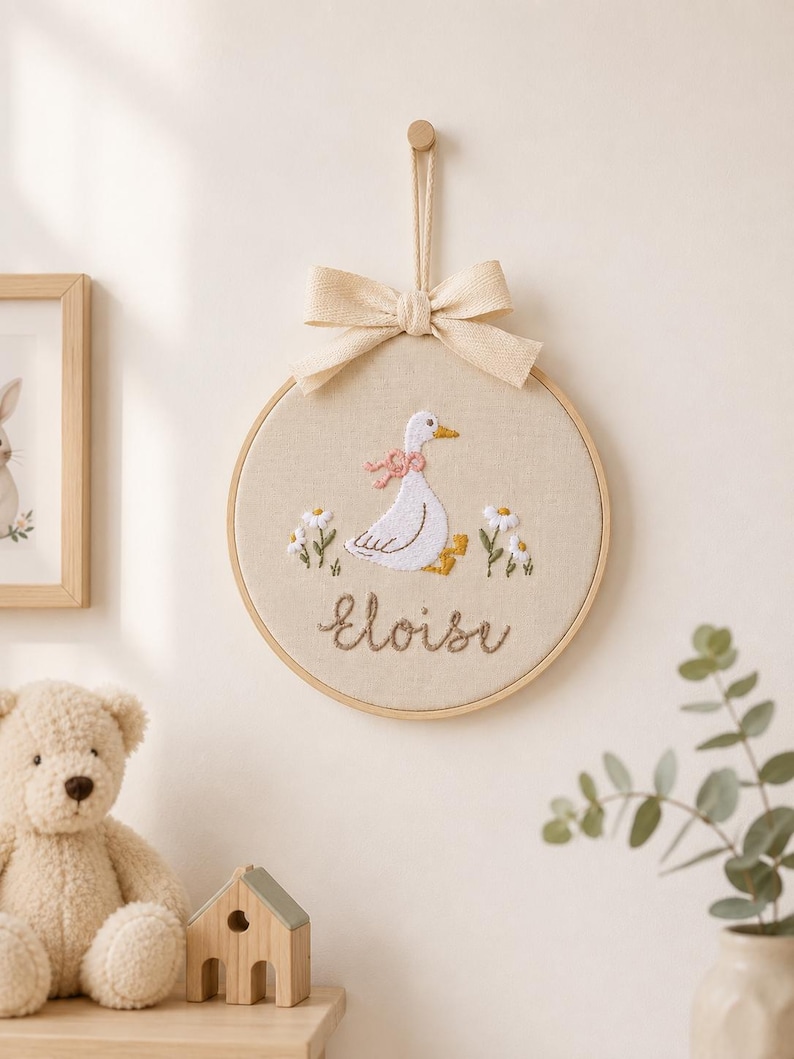 May include: A circular wooden embroidery hoop featuring a white goose with a pink neck ribbon, surrounded by small white flowers and green stems. The name Eloise is embroidered in brown script below the goose. A beige fabric bow hangs from the top.