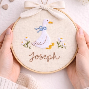 May include: A circular wooden embroidery hoop containing off-white fabric with a hand-stitched design of a white goose wearing a blue ribbon. The goose is surrounded by small white flowers with yellow centers. The name Joseph is stitched in brown.