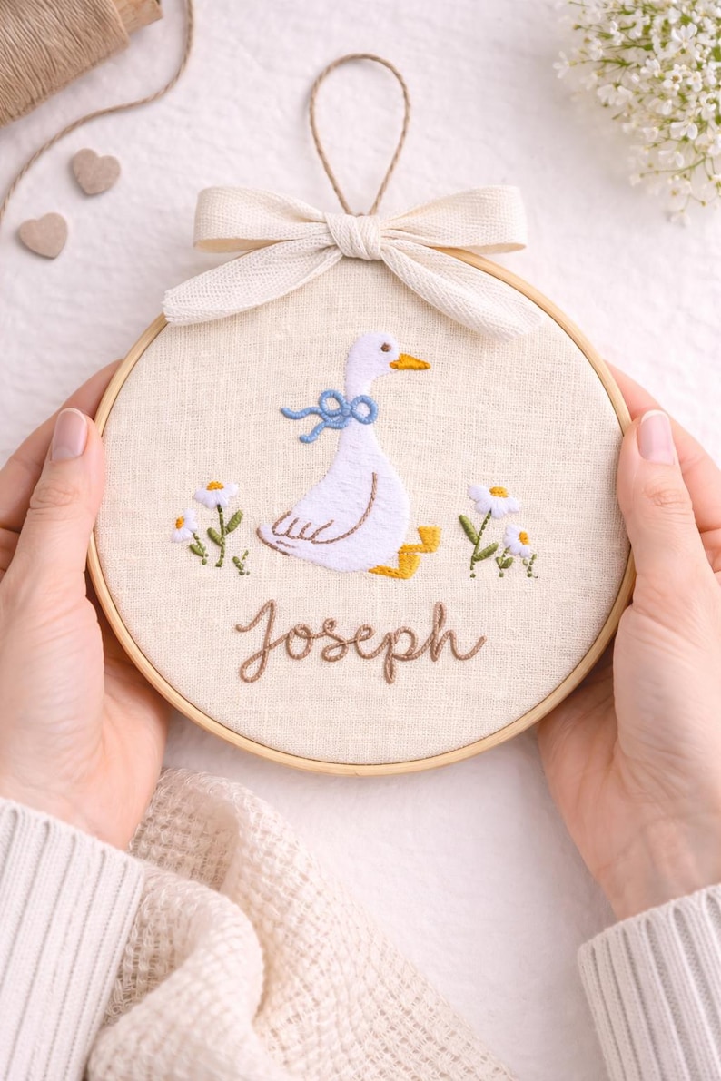 May include: A circular wooden embroidery hoop containing off-white fabric with a hand-stitched design of a white goose wearing a blue ribbon. The goose is surrounded by small white flowers with yellow centers. The name Joseph is stitched in brown.