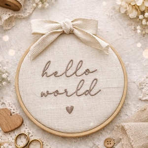 Op de afbeelding: Een beige borduurring met de woorden "hello world" geborduurd in bruin garen, een klein hartje en een beige strik. De ring is omgeven door kant, droogbloemen en naaigerei.