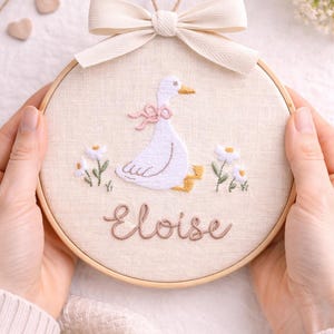May include: A circular wooden embroidery hoop containing a cream-colored fabric panel. The fabric features a hand-stitched white goose with a pink bow, yellow feet, and two white flowers. The name Eloise is embroidered in brown script below.