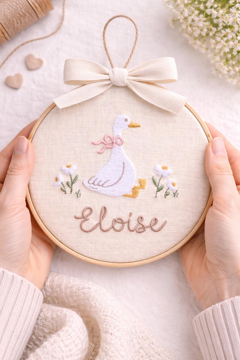 May include: A circular wooden embroidery hoop containing a cream-colored fabric panel. The fabric features a hand-stitched white goose with a pink bow, yellow feet, and two white flowers. The name Eloise is embroidered in brown script below.