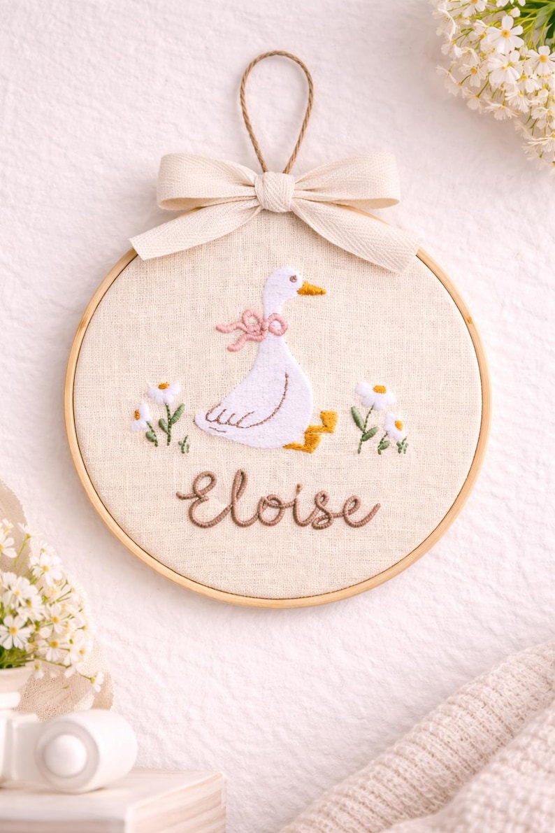 May include: A circular wooden embroidery hoop containing a beige fabric panel with a hand-stitched white goose wearing a pink bow. The goose is surrounded by small white flowers with yellow centers. The name Eloise is embroidered in brown script below.