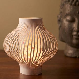 May include: A white paper lantern with a unique, curved design. The lantern's exterior features vertical, evenly spaced lines, while the interior glows with a soft light and a pattern of hearts. The lamp is a decorative home accent.