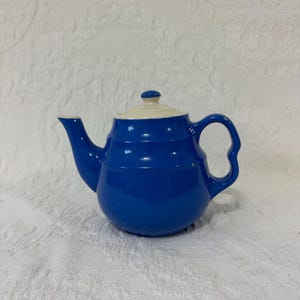May include: A glossy cobalt blue ceramic teapot featuring a rounded body with horizontal ridges, a curved handle, a pointed spout, and a removable lid with a cream-colored top section and a small blue knob handle on the center of the lid.