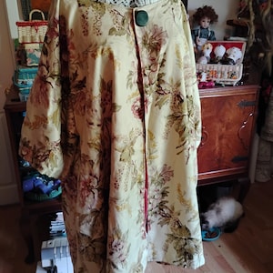 May include: A cream-coloured jacket featuring a floral pattern in shades of brown, green, and muted pink. The garment has a wide, loose silhouette, three-quarter length sleeves, and a single large, dark green circular button at the neckline.