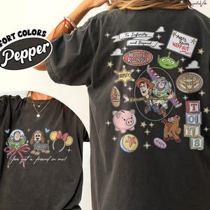 May include: Pepper-colored t-shirt featuring Toy Story characters and phrases. The front displays a graphic with Buzz Lightyear, a dog, and Minnie Mouse ears. The back showcases Woody, Buzz, and other characters with the text "To Infinity and Beyond."