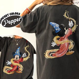 May include: Dark gray Comfort Colors t-shirt featuring a graphic of Mickey Mouse as a sorcerer, wearing a blue hat with stars and a red robe. The design includes a golden magic wand and swirling golden accents. The text "Comfort Colors Pepper" is on the upper left.