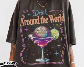 May include: A charcoal gray short-sleeve t-shirt featuring a graphic print of a cocktail glass with a geodesic dome sphere inside. The text reads Drink Around the World, surrounded by colorful stars and a lime slice against a purple border.
