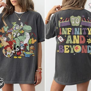 Op de afbeelding: Donkergrijs t-shirt met een kleurrijke afbeelding van Toy Story-personages op de voorkant. De achterkant van het shirt heeft de tekst "To Infinity and Beyond" in een retro lettertype. Het shirt is gelabeld "Comfort Colors Pepper".