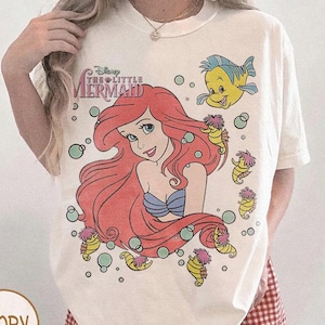 May include: Ivory-colored t-shirt featuring a graphic of Ariel from Disney's The Little Mermaid. The design includes Ariel with red hair, a blue top, and a yellow fish, surrounded by seahorses and bubbles. The text "Disney The Little Mermaid" is above Ariel.