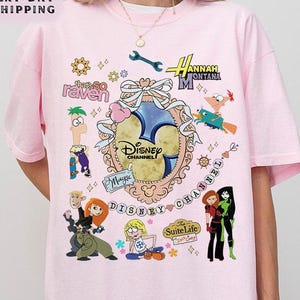 Könnte beinhalten: Hellrosa T-Shirt mit verschiedenen Disney Channel Cartoon-Charakteren und Logos. Das Shirt enthält den Text "Disney Channel" und "Hannah Montana". Das Design ist farbenfroh und nostalgisch, perfekt für Fans des Senders.