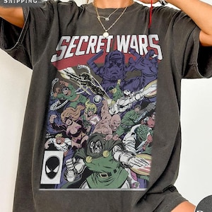 Puede incluir: Camiseta gris oscuro con una ilustraci&oacute;n de c&oacute;mic que dice "SECRET WARS". El gr&aacute;fico incluye varios personajes en colores vibrantes, con una pancarta roja y blanca en la parte superior. Se ve un logo de Spider-Man.