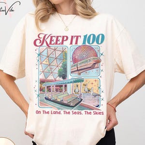 May include: Ivory-colored t-shirt with a colorful graphic design. The design includes the text "KEEP IT 100" and illustrations of a geometric structure, a monorail, and a boat with ducks. The text "On The Land, The Seas, The Skies" is also included.
