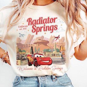 Puede incluir: Camiseta color crema con un gr&aacute;fico de Radiator Springs de la pel&iacute;cula Cars. El dise&ntilde;o incluye el texto "Radiator Springs California est. 2012" y "Welcome to Radiator Springs" con una imagen de Rayo McQueen.