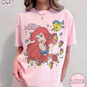 May include: A light pink t-shirt featuring a graphic of Ariel from Disney's The Little Mermaid. The design includes Ariel with red hair, a blue top, and Flounder. The shirt also has the movie's logo and seahorses.