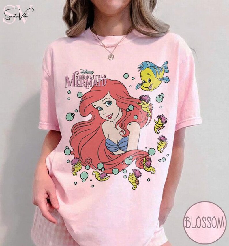May include: A light pink t-shirt featuring a graphic of Ariel from Disney's The Little Mermaid. The design includes Ariel with red hair, a blue top, and Flounder. The shirt also has the movie's logo and seahorses.