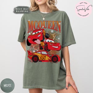 K&ouml;nnte beinhalten: Ein moosgr&uuml;nes T-Shirt mit einer Grafik der Figur Lightning McQueen aus dem Film Cars. Das Design zeigt das Wort "McQueen" in Orange und Rot, mit Bildern des Autos. Die Nummer "95" ist ebenfalls sichtbar.