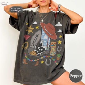 May include: A charcoal gray short-sleeve t-shirt featuring a graphic print of a cowboy boot with cow print, a brown cowboy hat, a sheriff star, alphabet blocks with the letters T, S, and 13, a lasso, and a colorful beaded string with text.