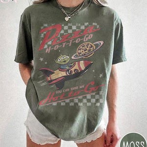 May include: Moss green t-shirt with a graphic featuring a cartoon alien in a rocket ship, a pizza planet, and the text "Pizza H-O-T-T-O-Go". The shirt has a vintage, distressed look.
