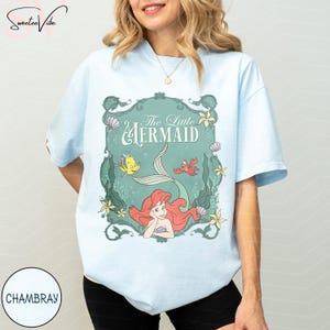 May include: Light blue t-shirt featuring a graphic from "The Little Mermaid." The design includes Ariel, Flounder, Sebastian, and the film's title in a decorative frame. The shirt is a soft, casual style.
