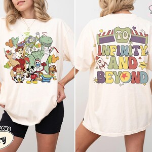 Op de afbeelding: Ivoor kleurig T-shirt met een kleurrijke afbeelding van animatiefiguren, waaronder een groene dinosaurus, Woody, Buzz Lightyear en Mickey Mouse. De achterkant van het shirt toont de zin "TO INFINITY AND BEYOND" in een retro lettertype, met een raket en sterren.