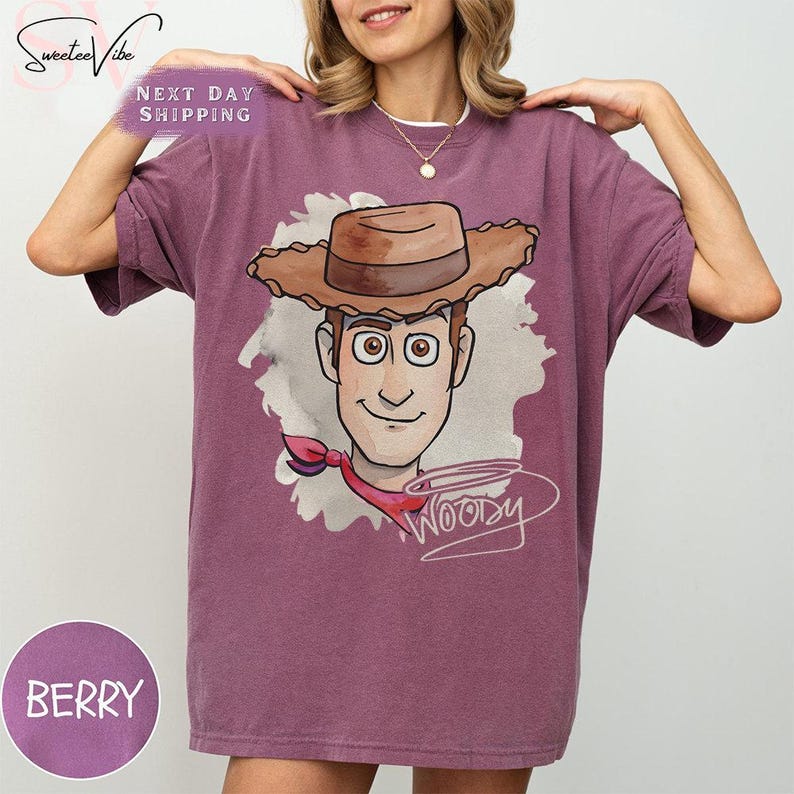 Puede incluir: Camiseta de color bayas con una ilustraci&oacute;n de dibujos animados de Woody de Toy Story. El dise&ntilde;o incluye la cara de Woody, su sombrero de vaquero y su firma. La camiseta tiene un corte holgado y mangas cortas. El texto "Woody" est&aacute; escrito en cursiva.