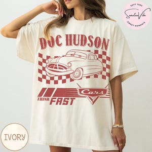 Puede incluir: Camiseta de color marfil con un gr&aacute;fico rojo de Doc Hudson de la pel&iacute;cula Cars, con el texto "DOC HUDSON" encima del coche y "THINK FAST" debajo. El dise&ntilde;o incluye un fondo a cuadros y el logotipo de Cars.