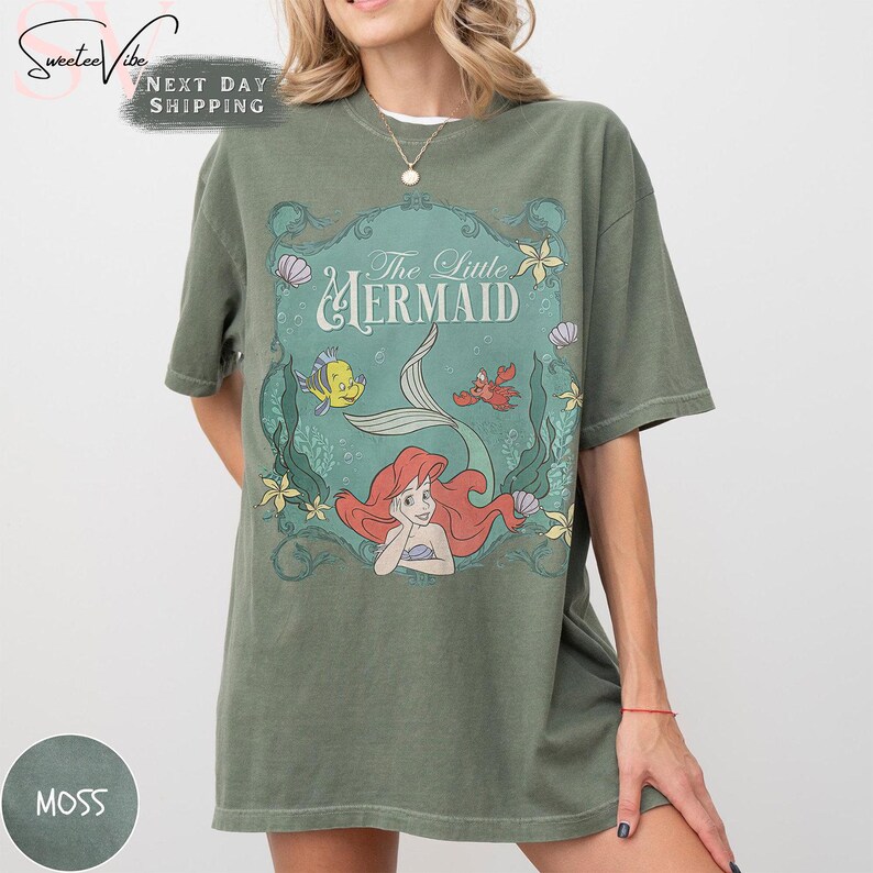 May include: Moss green t-shirt featuring a graphic from Disney's The Little Mermaid. The design includes Ariel, Flounder, Sebastian, and the words "The Little Mermaid". The shirt has short sleeves and a crew neck.