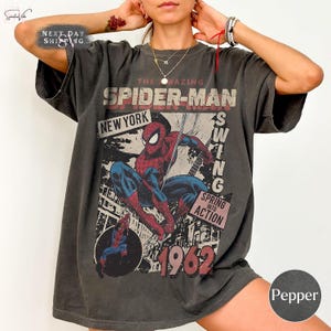 May include: A dark gray t-shirt featuring a vintage Spider-Man graphic. The design includes the words "The Amazing Spider-Man," "New York," and "1962." The superhero is depicted in red and blue, swinging into action.