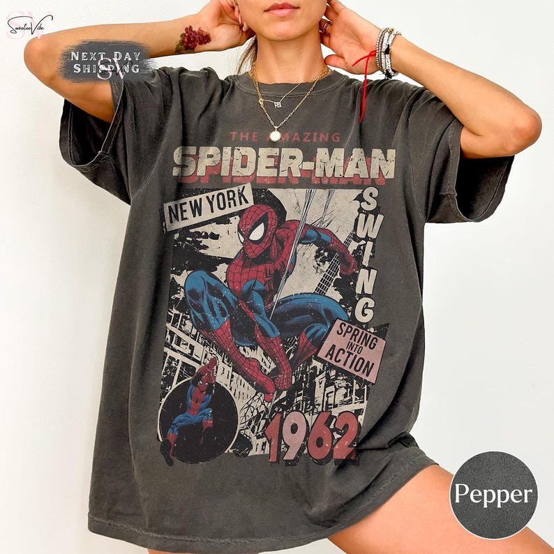 May include: A dark gray t-shirt featuring a vintage Spider-Man graphic. The design includes the words "The Amazing Spider-Man," "New York," and "1962." The superhero is depicted in red and blue, swinging into action.