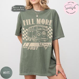 Puede incluir: Una camiseta verde musgo con un gr&aacute;fico de furgoneta retro y el texto "FILL MORE CARS THINK FAST". El dise&ntilde;o incluye una bandera a cuadros. La camiseta tiene un corte holgado y mangas cortas.