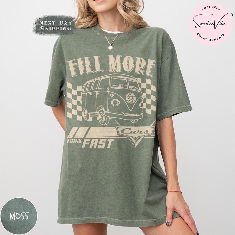 Puede incluir: Una camiseta verde musgo con un gr&aacute;fico de furgoneta retro y el texto "FILL MORE CARS THINK FAST". El dise&ntilde;o incluye una bandera a cuadros. La camiseta tiene un corte holgado y mangas cortas.