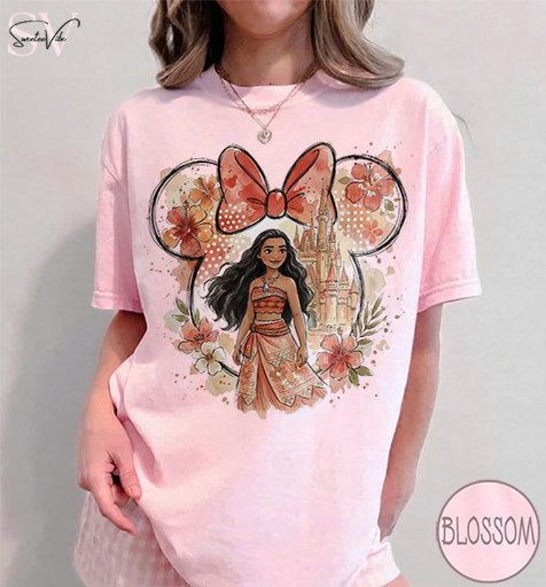 May include: Pink t-shirt featuring a Minnie Mouse-shaped design with a red bow, flowers, and a castle. The design includes a Moana character in a dress. The word "Blossom" is in a pink circle on the bottom right.