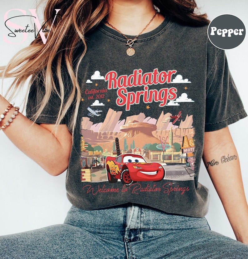 Puede incluir: Camiseta gris oscuro con un dise&ntilde;o de Rayo McQueen de la pel&iacute;cula Cars, y el texto "Radiator Springs" en rojo. El dise&ntilde;o incluye un paisaje des&eacute;rtico y las palabras "California est. 2012" y "Welcome to Radiator Springs."