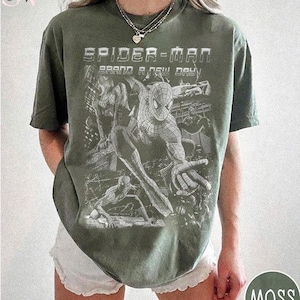 May include: An olive green short-sleeve t-shirt featuring a white graphic print of Spider-Man in various action poses. The text "SPIDER-MAN" and "BRAND A NEW DAY" is printed in a bold, distressed font above the character illustrations.
