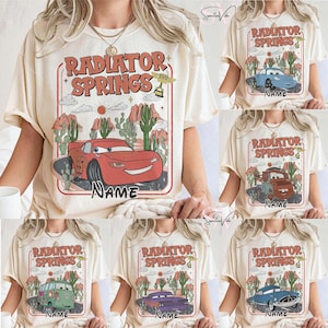 May include: Cream-colored t-shirts featuring a graphic design with the text "RADIATOR SPRINGS" in orange and red. The design includes a red race car, cacti, and desert scenery. The word "NAME" is below the car.