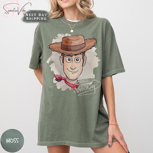 Puede incluir: Camiseta verde musgo con una ilustraci&oacute;n de dibujos animados de Woody de Toy Story. El dise&ntilde;o muestra la cara de Woody, un sombrero de vaquero marr&oacute;n y un pa&ntilde;uelo rojo. La palabra "Woody" est&aacute; escrita en cursiva. La camiseta es de corte holgado.