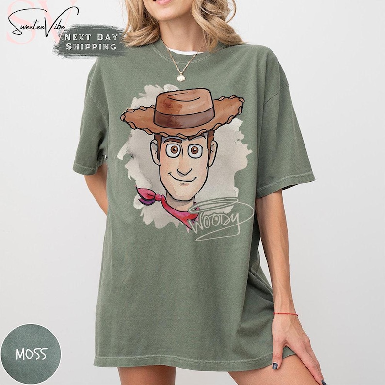 Puede incluir: Camiseta verde musgo con una ilustraci&oacute;n de dibujos animados de Woody de Toy Story. El dise&ntilde;o muestra la cara de Woody, un sombrero de vaquero marr&oacute;n y un pa&ntilde;uelo rojo. La palabra "Woody" est&aacute; escrita en cursiva. La camiseta es de corte holgado.