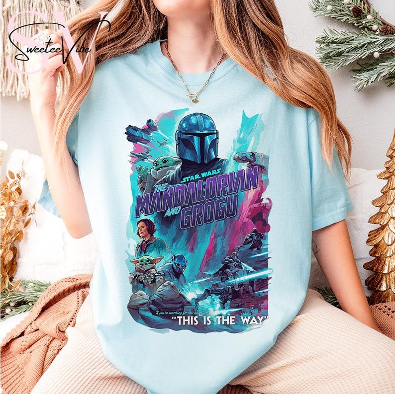 May include: Light blue t-shirt featuring a colorful graphic from the Star Wars series, "The Mandalorian and Grogu." The design includes the Mandalorian, Grogu, and other characters, with the phrase "THIS IS THE WAY" at the bottom.