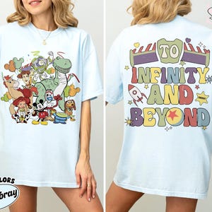 Op de afbeelding: Lichtblauw T-shirt met een kleurrijke afbeelding van Toy Story-personages op de voorkant. De achterkant van het shirt toont de zin "To Infinity and Beyond" in een retro lettertype. Het shirt is gemaakt van Comfort Colors Chambray stof.