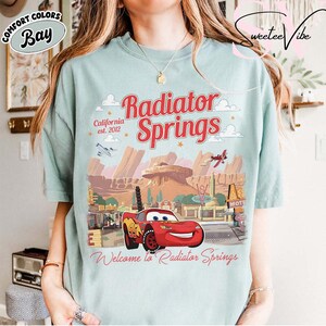 Puede incluir: Camiseta azul claro con un dise&ntilde;o de Radiator Springs de la pel&iacute;cula Cars. El dise&ntilde;o incluye el texto "Radiator Springs", "California est. 2012" y "Welcome to Radiator Springs". El dise&ntilde;o tambi&eacute;n presenta un coche de carreras rojo.
