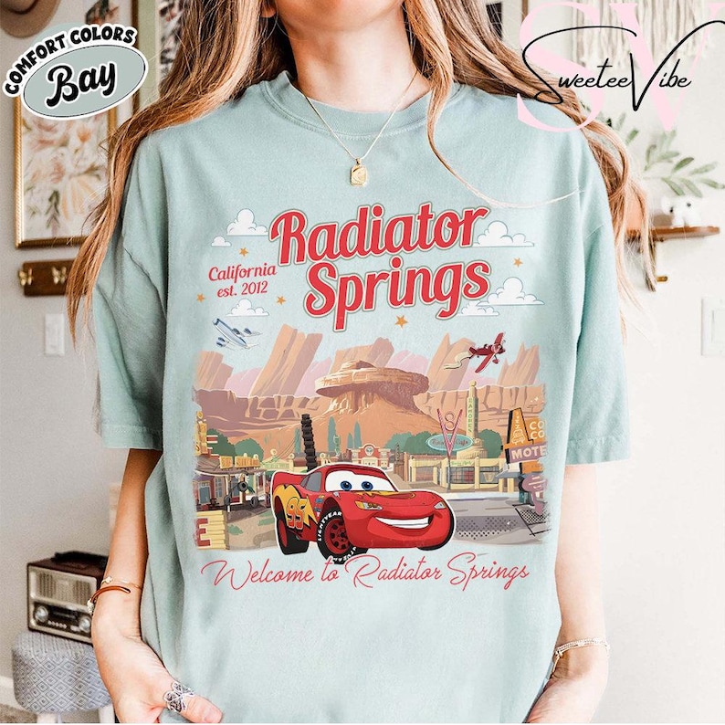 Puede incluir: Camiseta azul claro con un dise&ntilde;o de Radiator Springs de la pel&iacute;cula Cars. El dise&ntilde;o incluye el texto "Radiator Springs", "California est. 2012" y "Welcome to Radiator Springs". El dise&ntilde;o tambi&eacute;n presenta un coche de carreras rojo.
